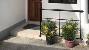 Um edifício branco com porta de madeira, janela com moldura de metal e guarda-corpo preto. Há duas plantas em vasos com flores amarelas na frente.