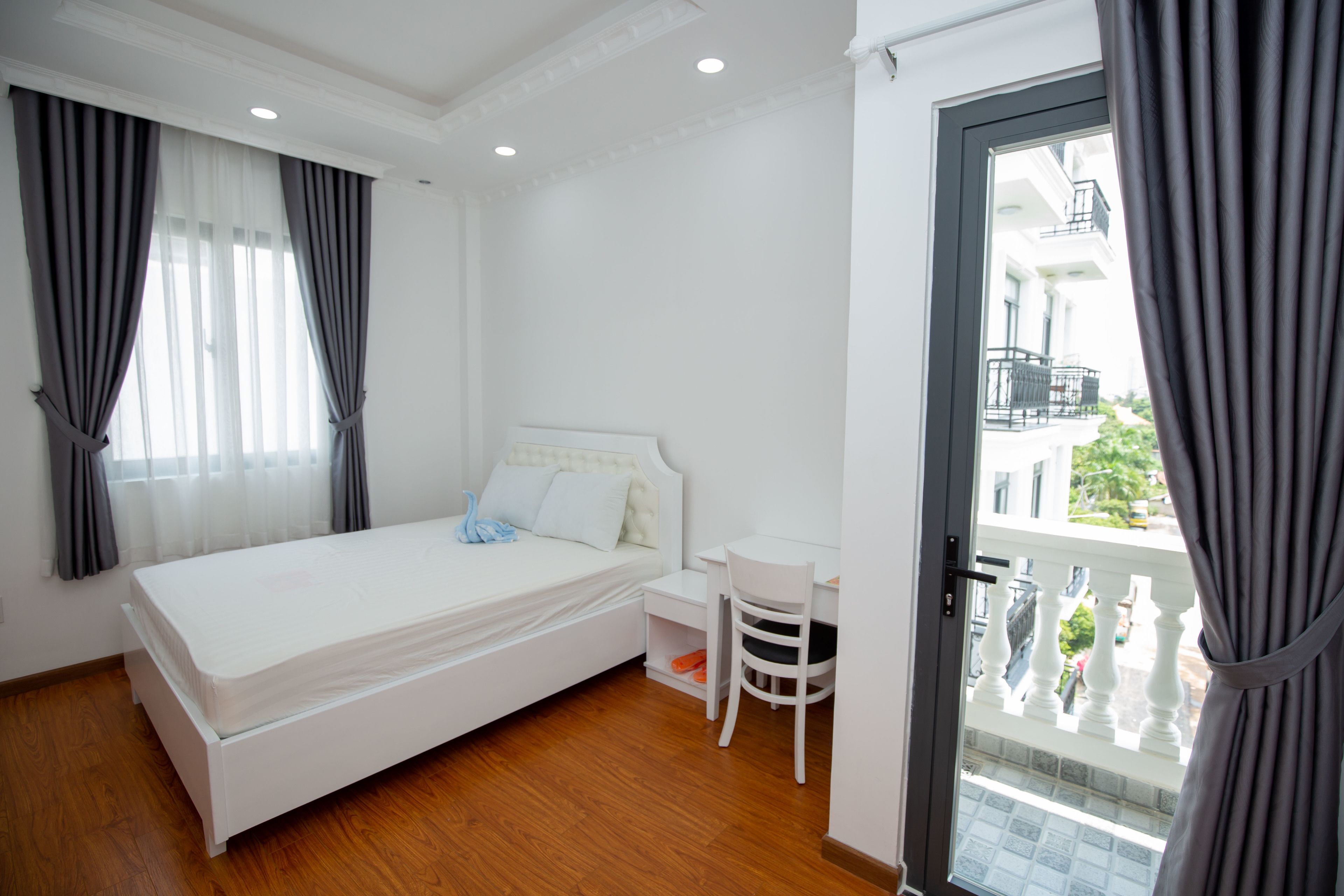 A bedroom with a bed, a desk, and a chair. There is a balcony with a railing and a view of buildings.