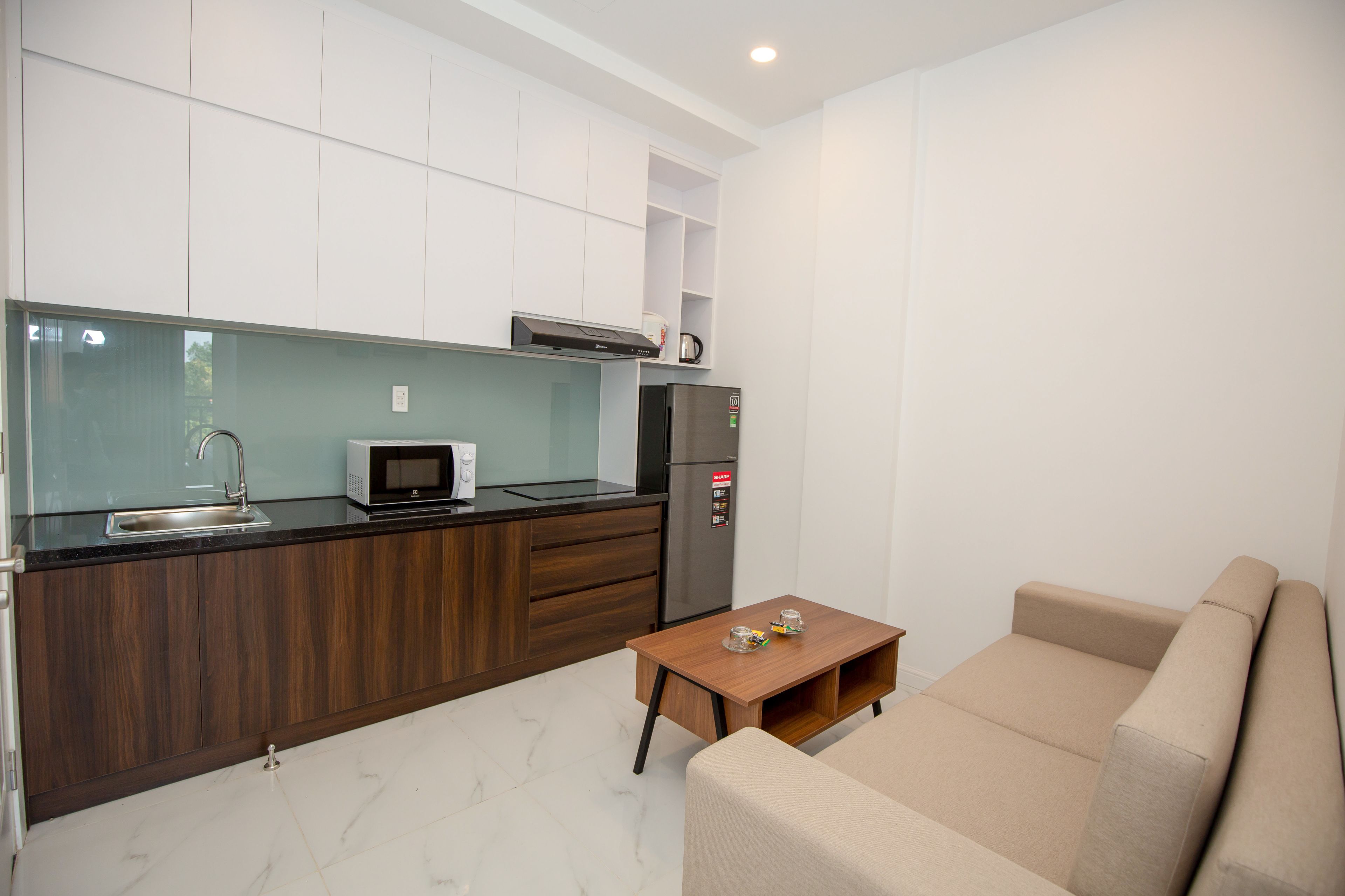 Una cocina moderna con microondas, refrigerador y armarios de madera. En la zona de estar, hay una mesa pequeña de madera y un sofá color beige.