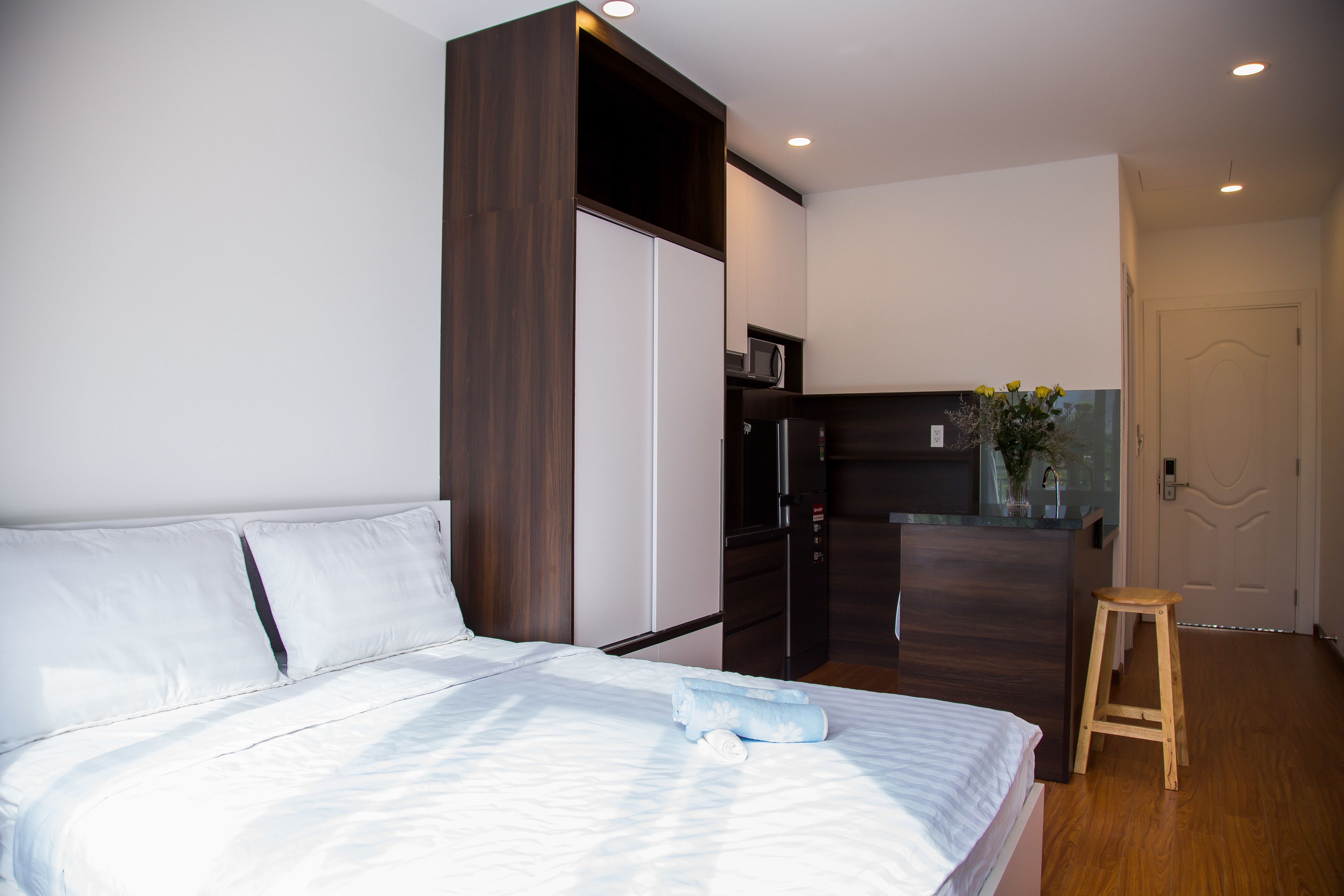 A bedroom with a bed, a wooden wardrobe, a small stool, and a kitchenette area with a microwave and refrigerator.