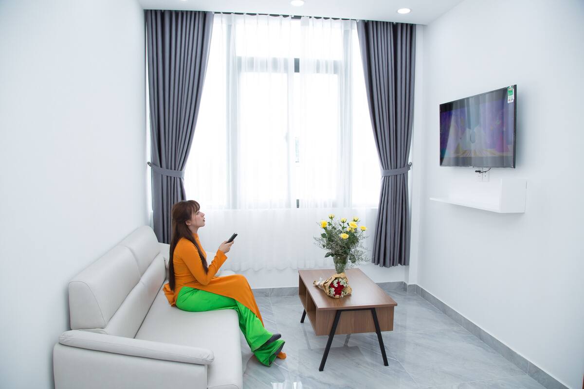 En moderne stue med sofa, sofabord med blomster, fladskærms-TV og store vinduer med gardiner.