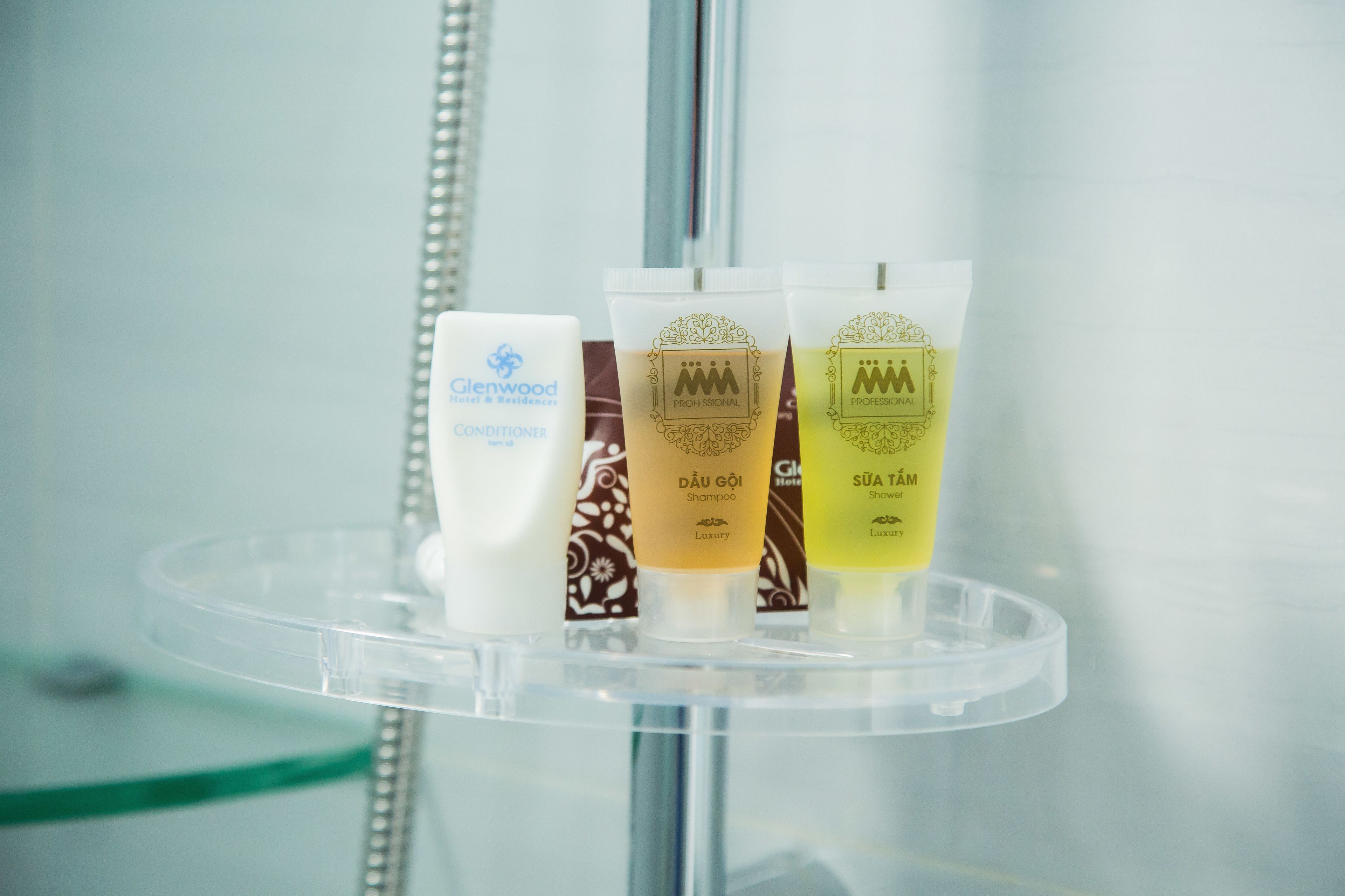 A bathroom shelf with three toiletries: a white bottle labeled "Glenwood Hotel & Residences," a brown bottle labeled "Dâu Gỏi Shampoo," and a yellow bottle labeled "Sửa Tâm Shower Gel."