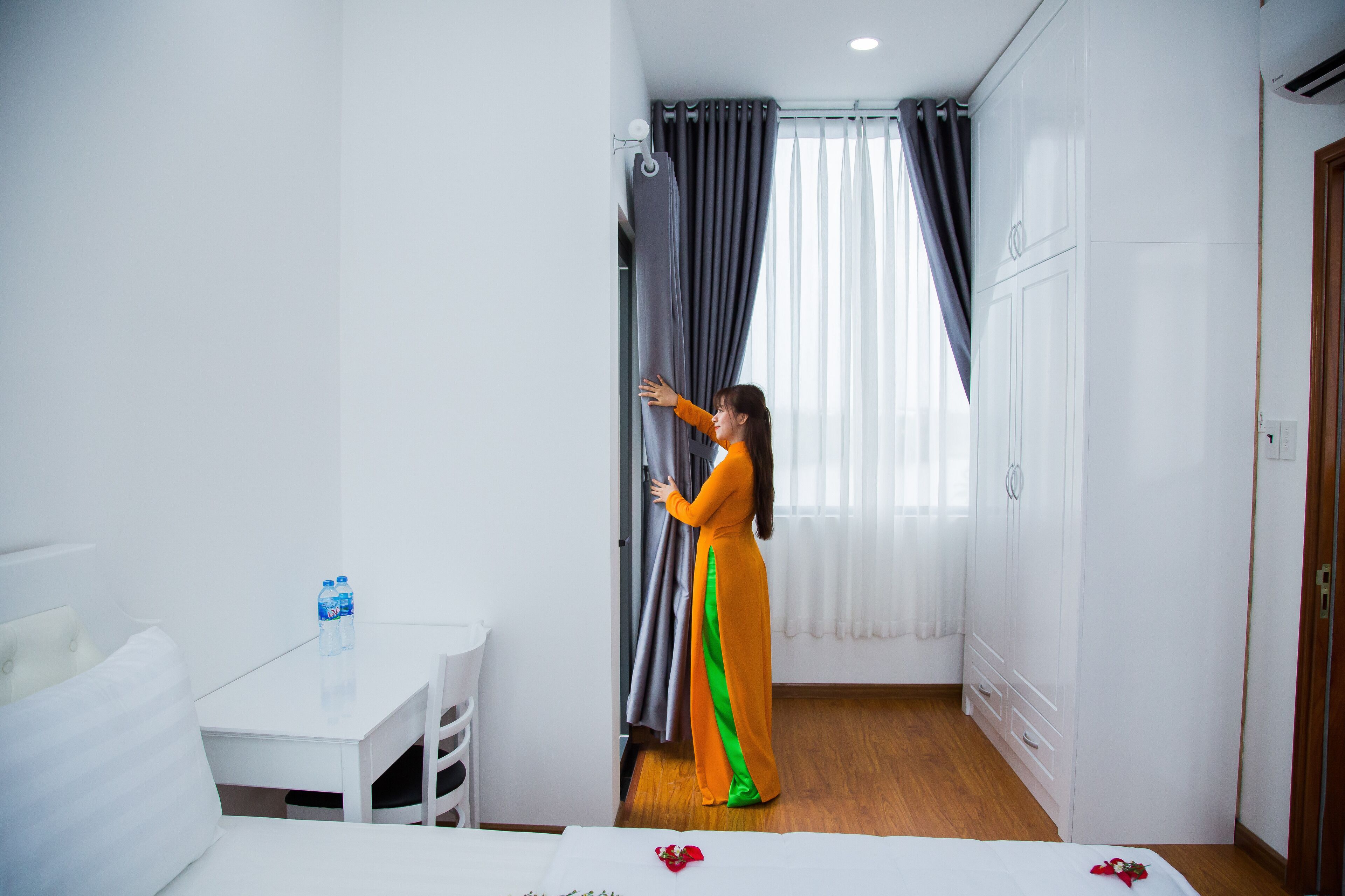 A room with a bed, a small table with a water bottle, and a person adjusting curtains.