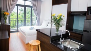 Ein modernes Hotelzimmer mit einem Bett, einem kleinen Holzschemel, einer Küchenzeile mit Spüle und einem großen Fenster mit Vorhängen.