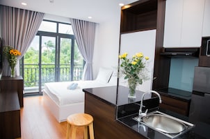 Una habitación de hotel moderna con una cama, un taburete de madera, una zona de cocina con un lavabo y un ventanal con cortinas.