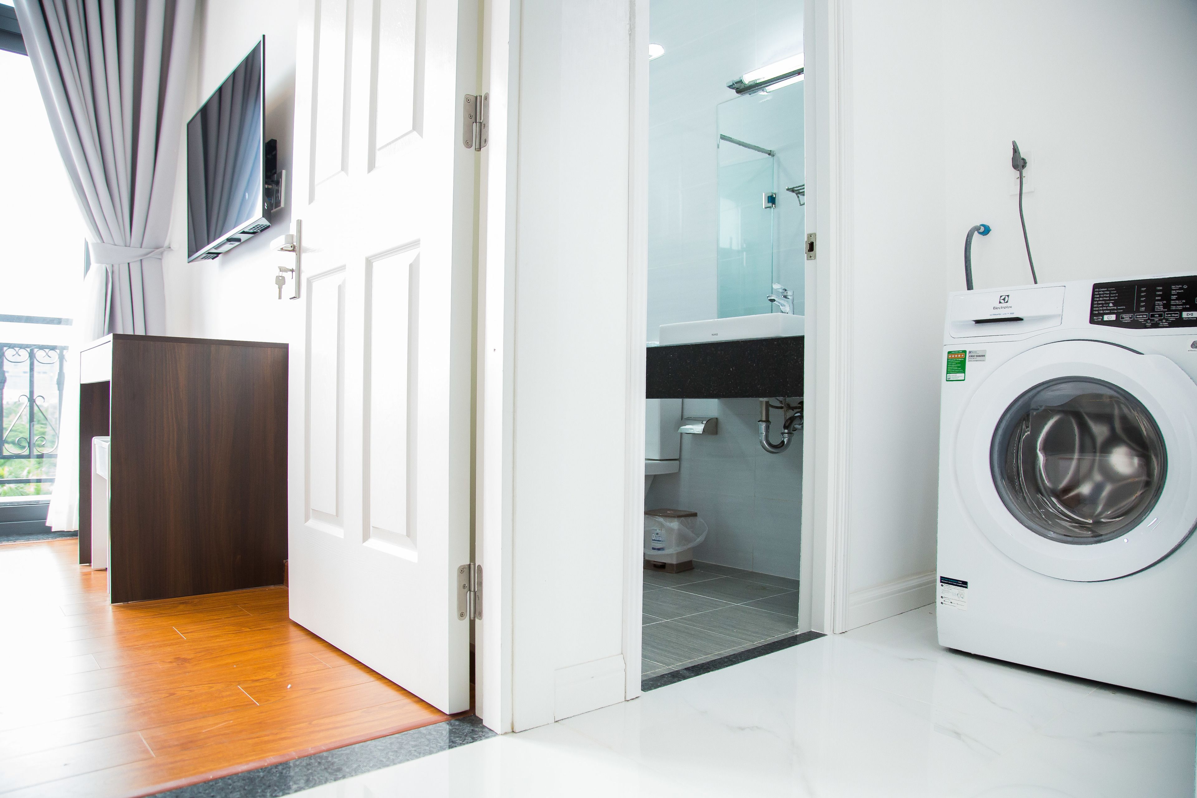 A modern hotel room with a wooden floor, a white door, a washing machine, a bathroom with a glass shower, and a mounted TV.