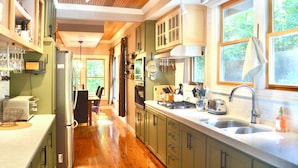 A kitchen with wooden flooring, green cabinets, a stainless steel sink, and a window with a view of greenery.