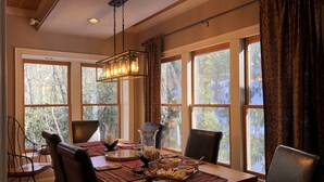 A dining area with a wooden table set for four, surrounded by chairs, with large windows offering a view of trees.