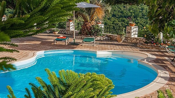 A swimming pool with a curved edge, surrounded by a stone deck and potted plants. - Villa Fantasia (Kefalonia)
