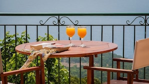 A round wooden table with two glasses of orange juice, a bowl, and a book on a balcony overlooking a scenic view. - Villa Fantasia (Kefalonia)