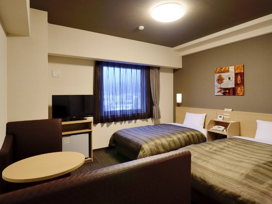 A hotel room with two beds, a desk with a television, a small table, and a window with curtains.