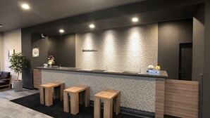 A reception area with a dark wall, a tiled backsplash, and a reception desk. There are three wooden stools and a potted plant. - Hotel Route-Inn Kashiwa Minami - Kokudo 16 gou zoi (Kashiwa)