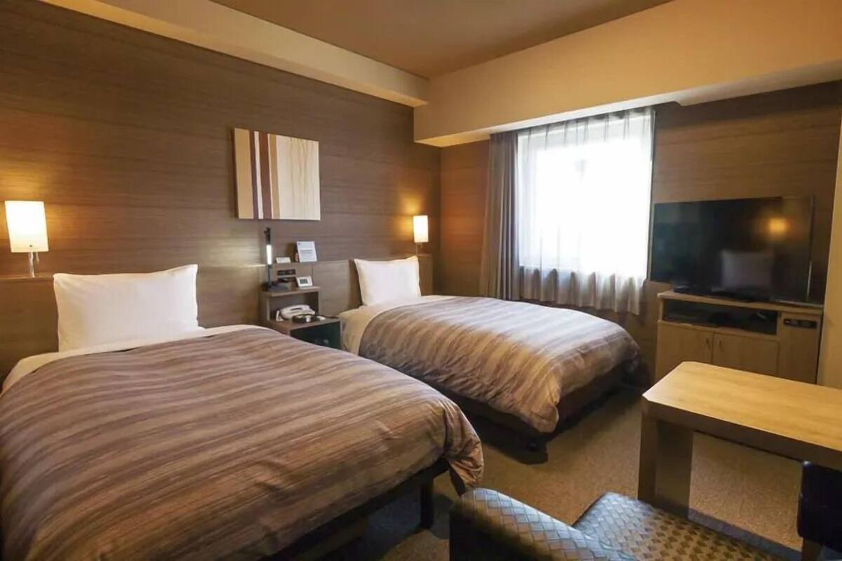 A hotel room with two beds, a TV, a desk, and a window with curtains.