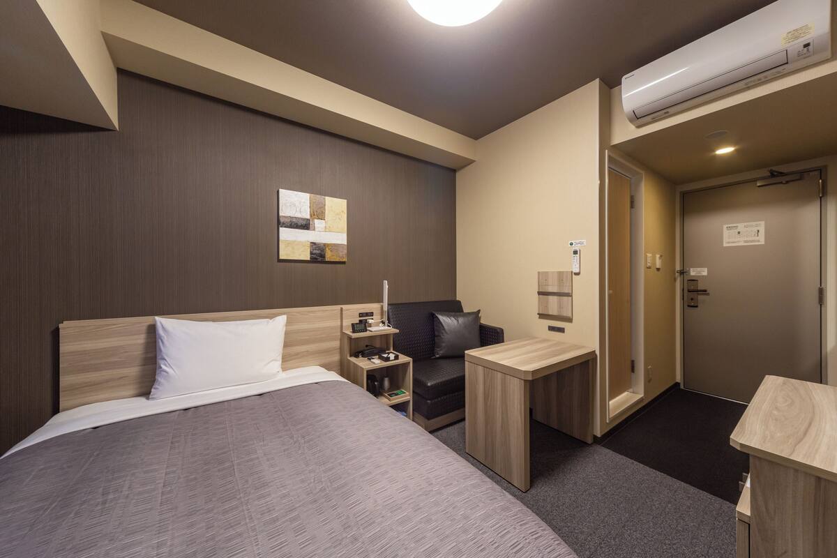 A hotel room with a large bed, a desk, a chair, a small table, and a wardrobe.
