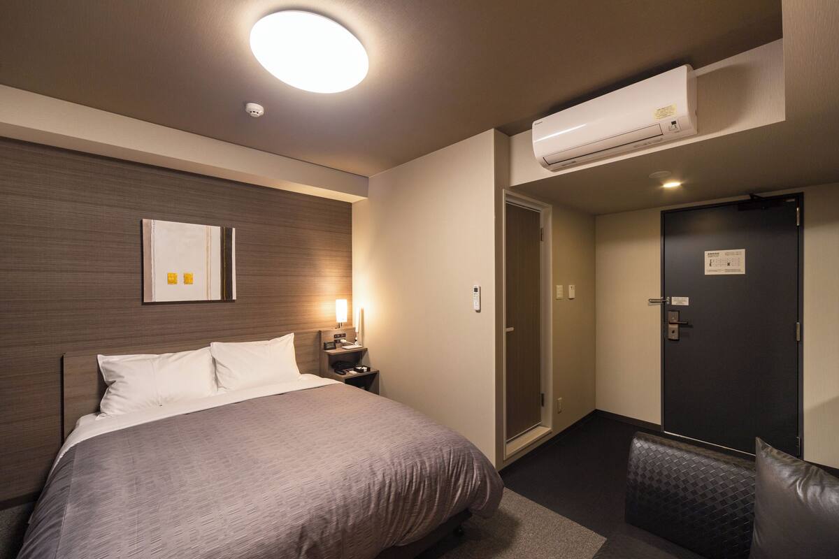 A hotel room with a bed, bedside table, lamp, air conditioning unit, and a door.