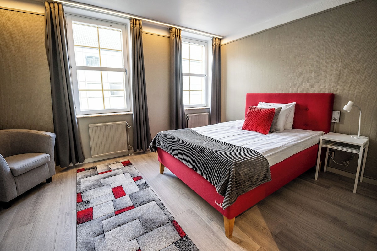 Une chambre à coucher avec un lit rouge, un tapis gris et rouge, un fauteuil gris et une table de chevet blanche avec une lampe.
