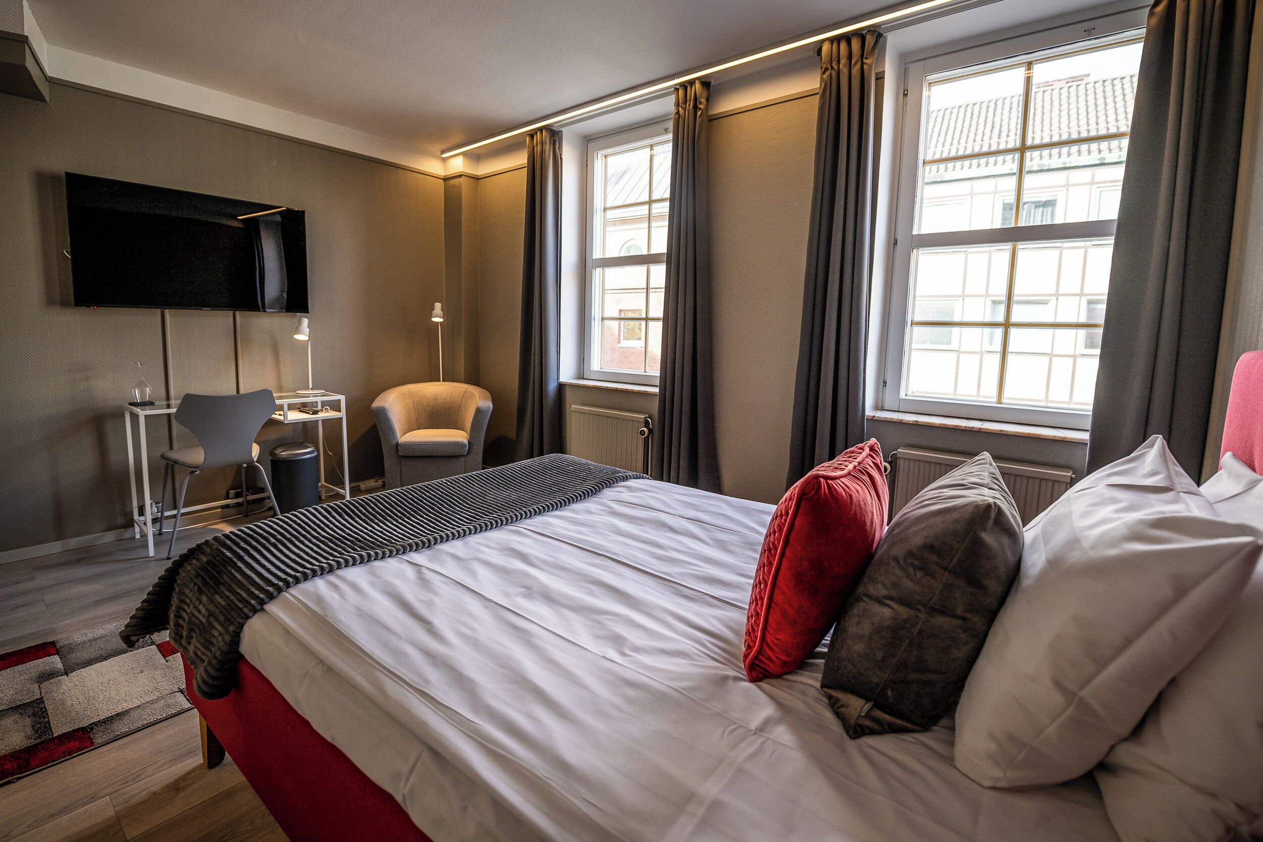 A hotel room with a bed, a desk, a chair, a TV, and large windows.