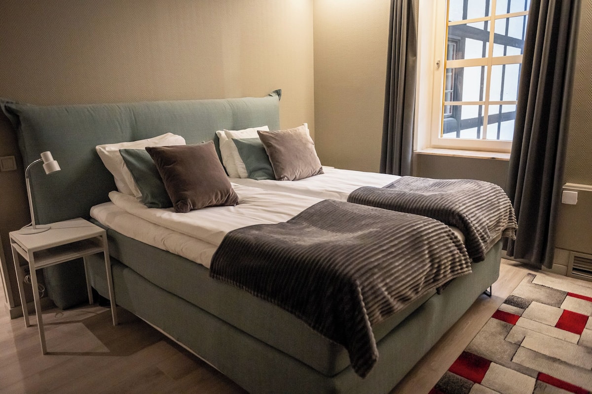 Un lit bien fait, avec une tête de lit verte, une table de chevet avec une lampe et une fenêtre avec des rideaux.