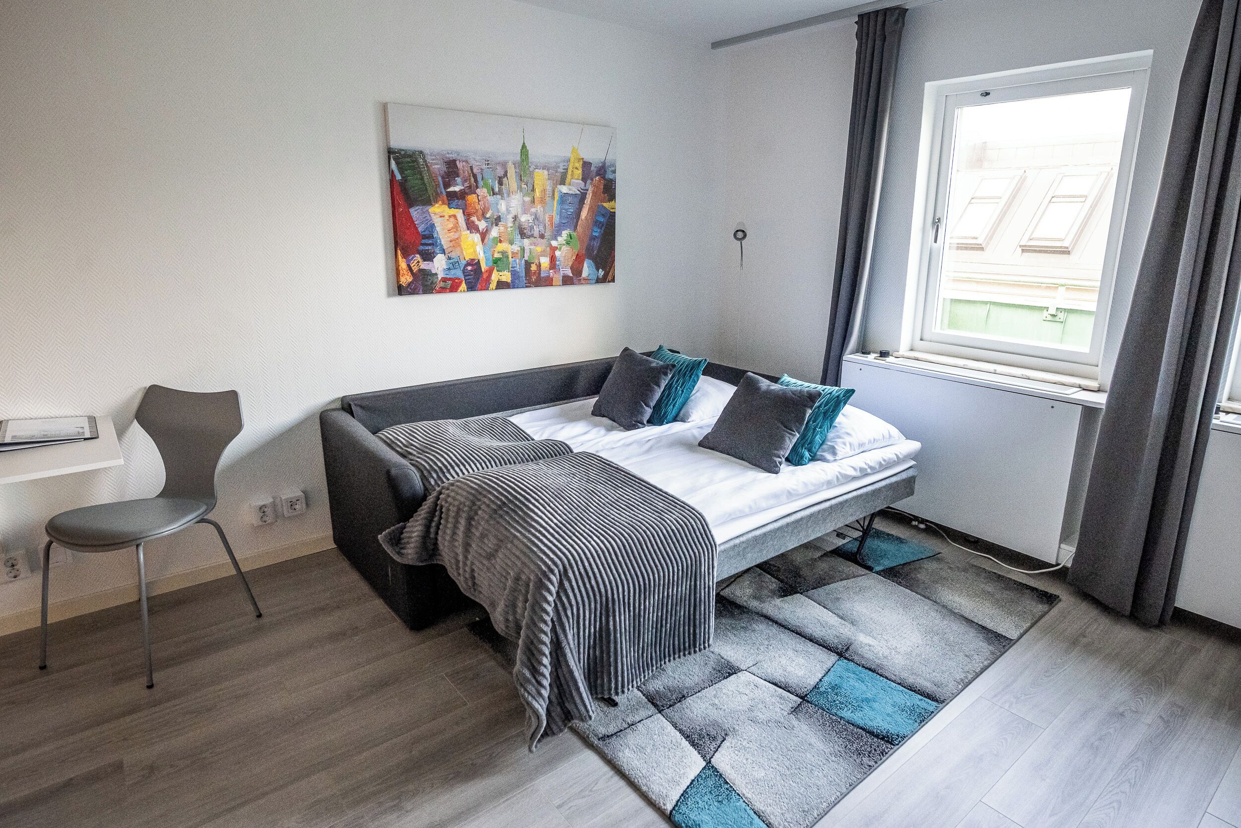 A modern bedroom with a bed, a chair, a cityscape painting, and a window with curtains.