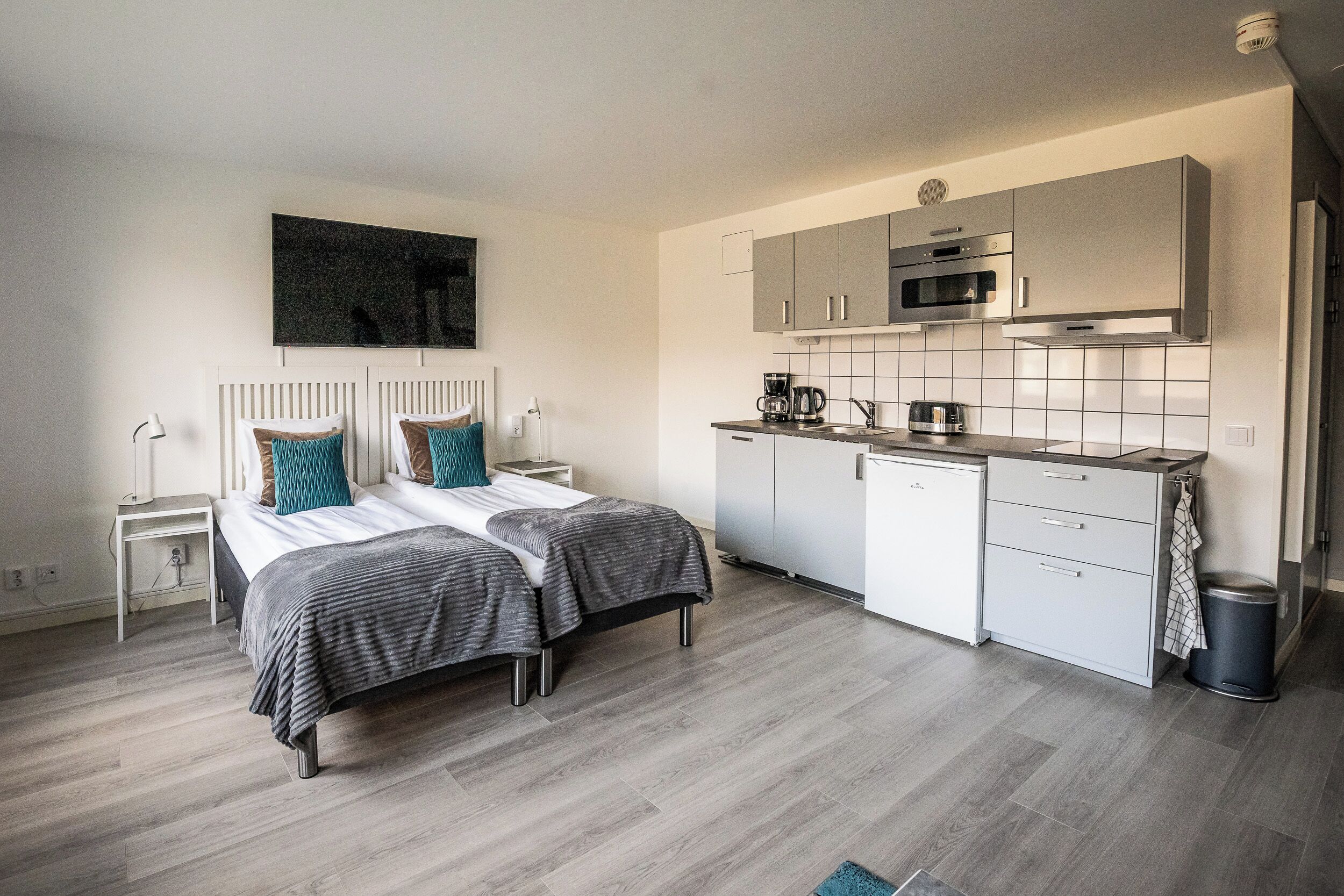 A modern bedroom with two beds, a small bedside table, and a nightstand. Adjacent is a compact kitchenette with a microwave, coffee maker, and a small refrigerator.