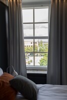 A room with a bed, a window with curtains, and a view of a building and trees outside.