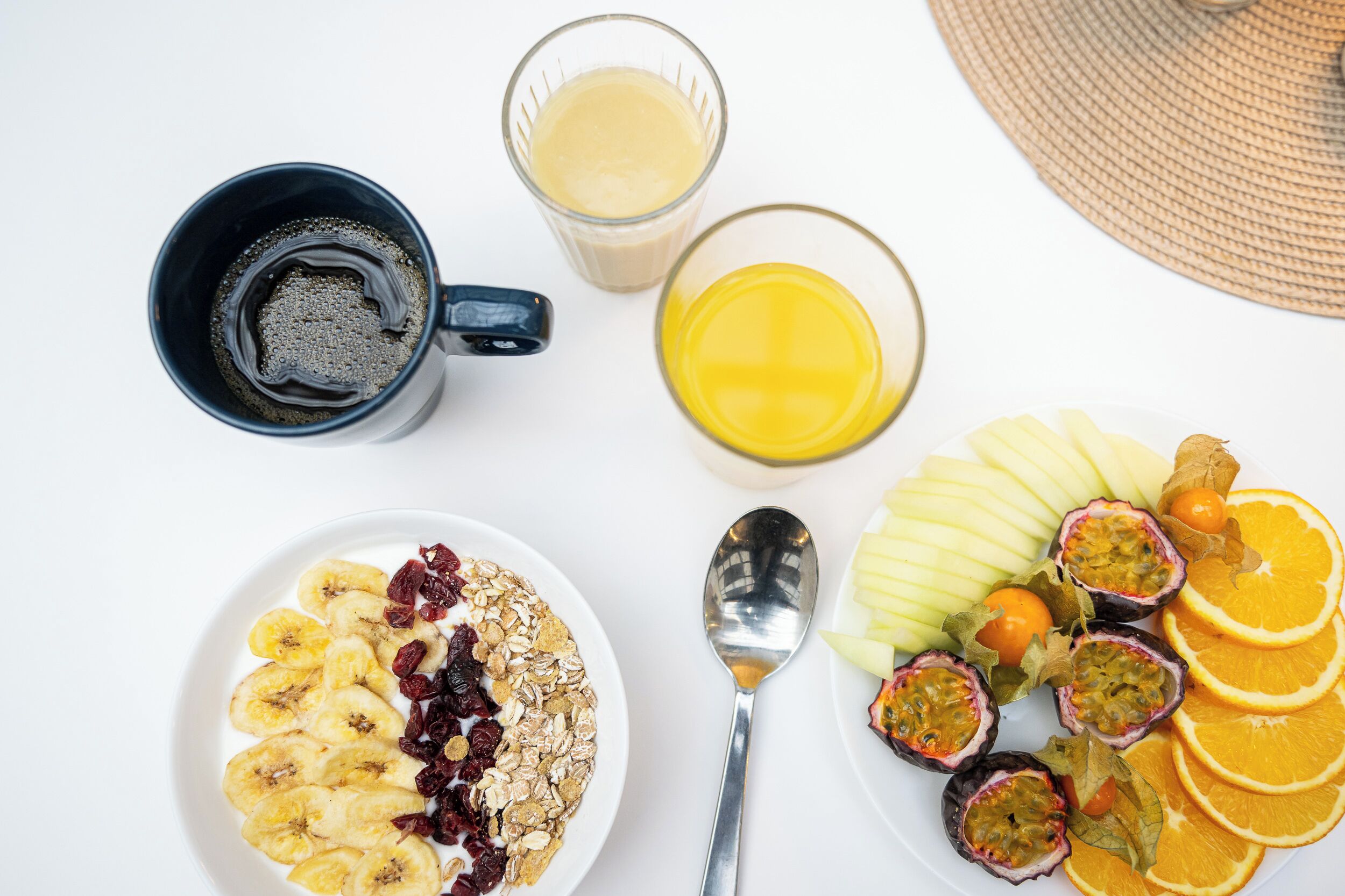 A breakfast setting with a plate of cereal, a bowl of yogurt, a cup of coffee, a glass of orange juice, and a plate of sliced fruits.