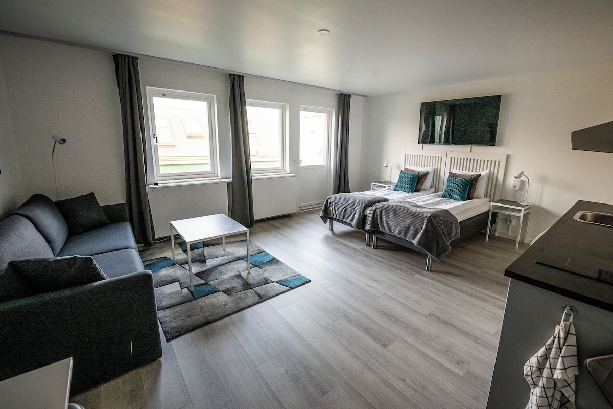 Une chambre d’hôtel moderne, dotée d’un canapé, d’un lit avec une tête de lit, d’une petite table et offrant une vue sur l’extérieur grâce à de grandes fenêtres.