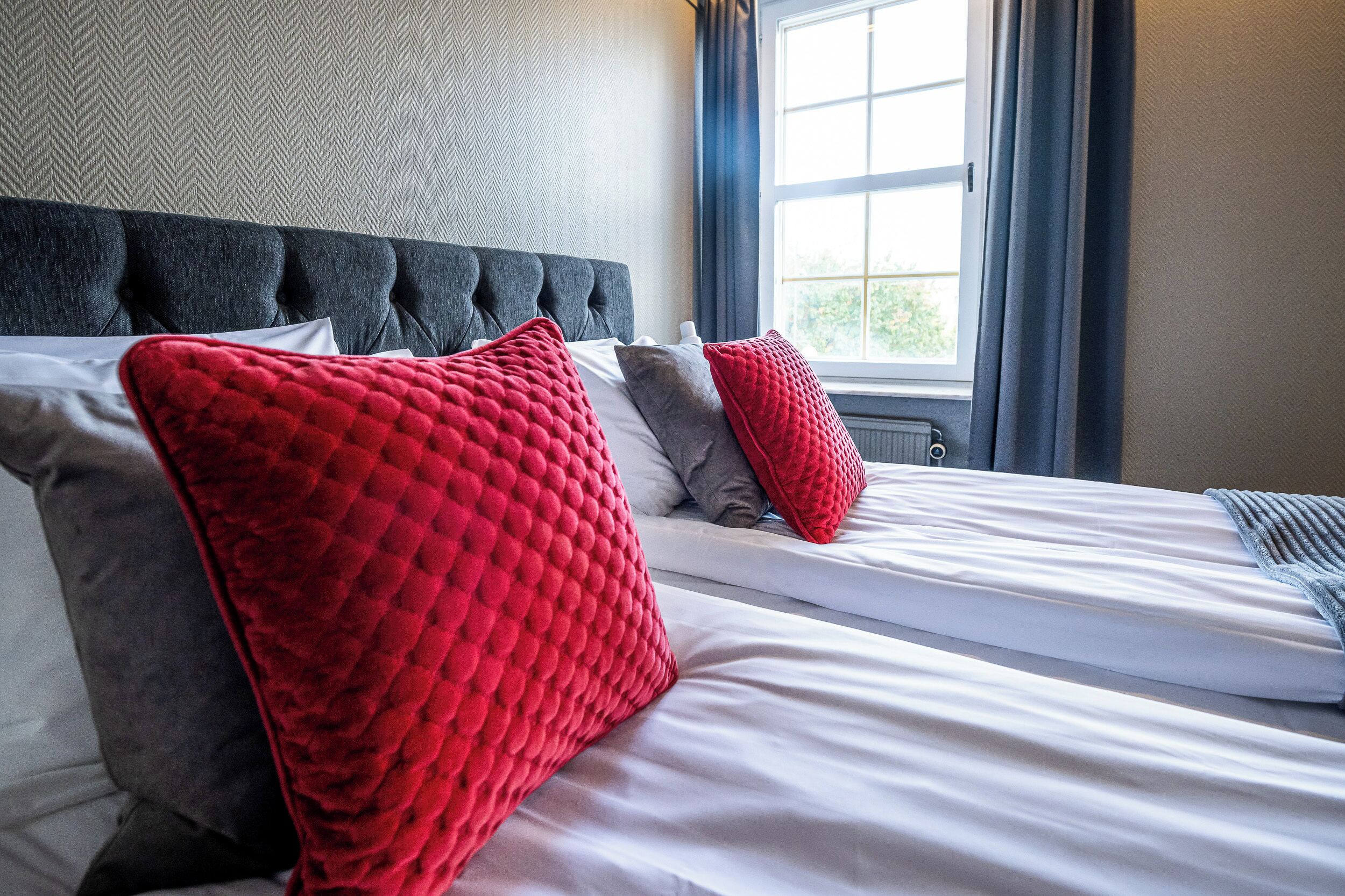A neatly made bed with white linens and red and grey pillows, a dark upholstered headboard, and a window with curtains.