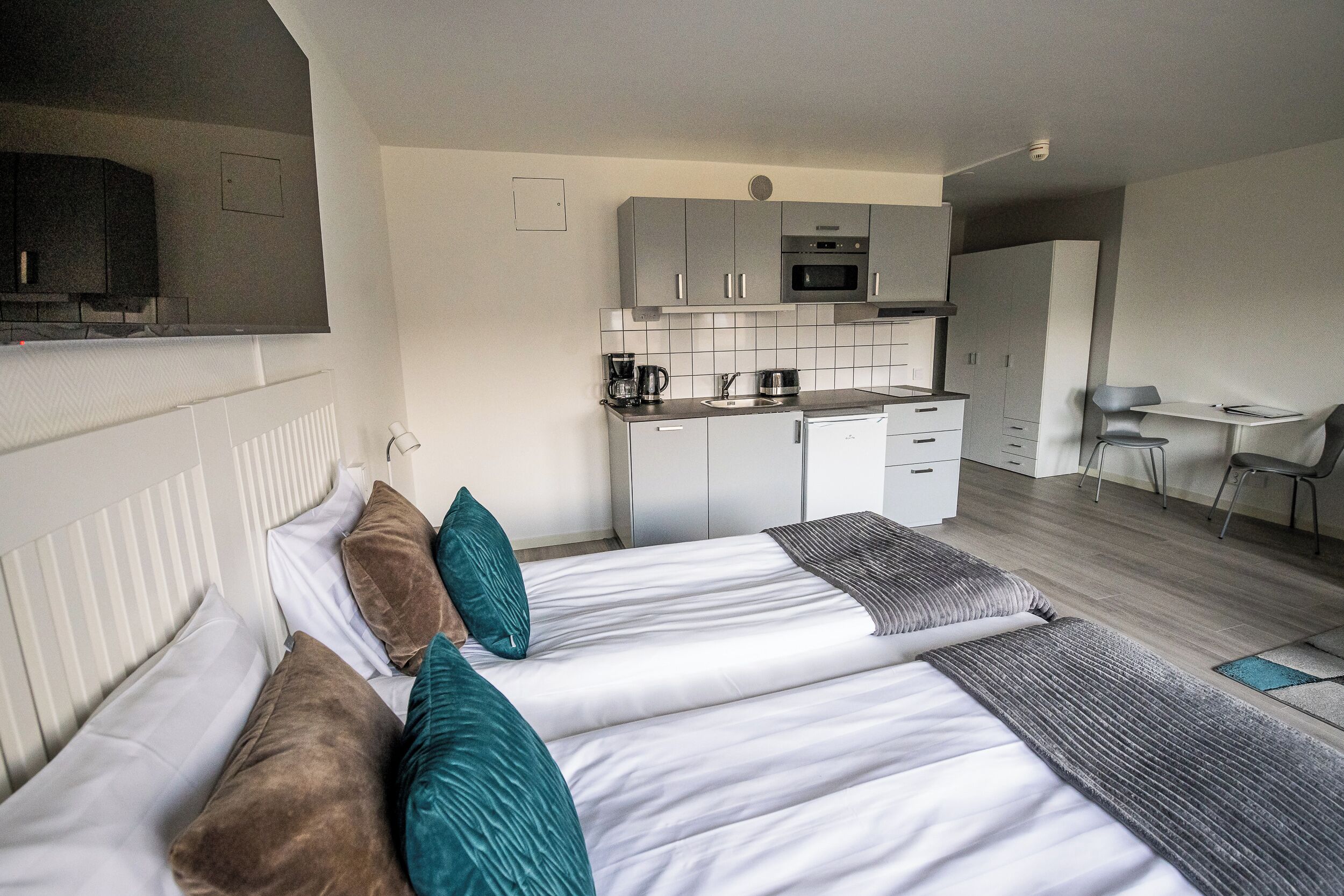 A modern hotel room with a bed, a small kitchenette, a desk, and a chair.