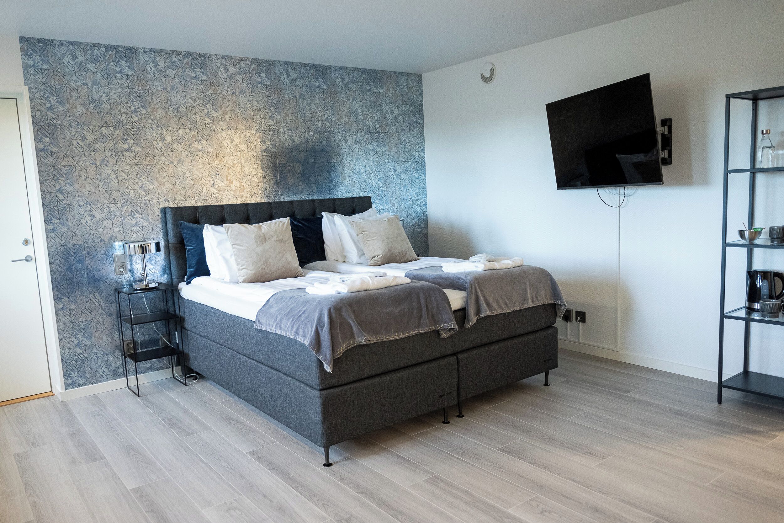 A modern bedroom with a grey bed, a TV mounted on the wall, and a small side table.