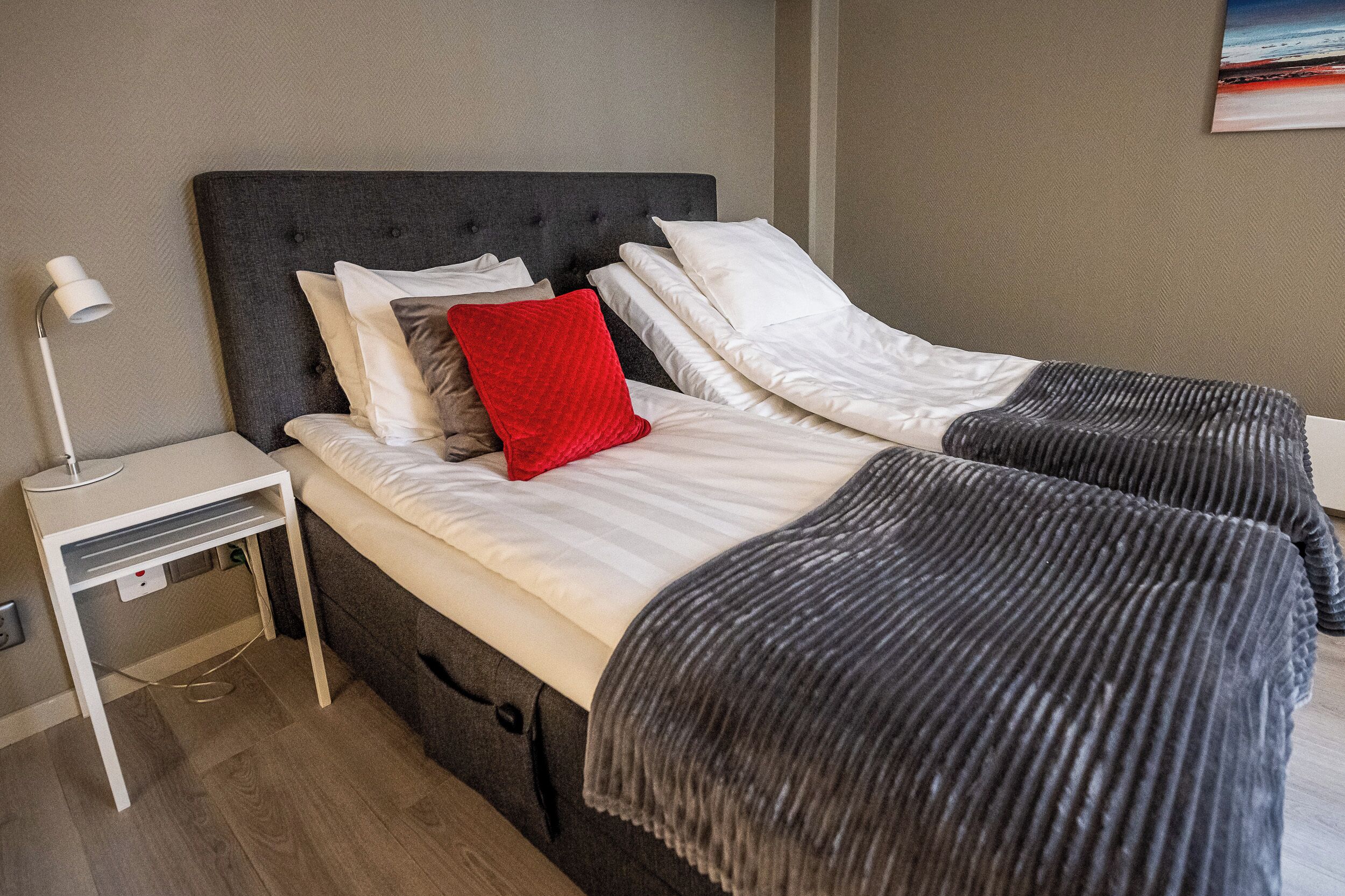 Een tweepersoonsbed met een grijs hoofdbord, wit beddengoed en een rood kussen. Er staat een nachtkastje met een lamp en een stopcontact.
