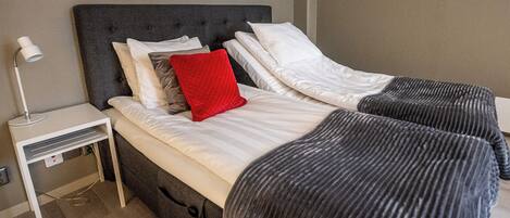Una cama doble con cabecera gris, ropa de cama blanca y una almohada roja. Hay una mesita de noche con una lámpara y un enchufe.