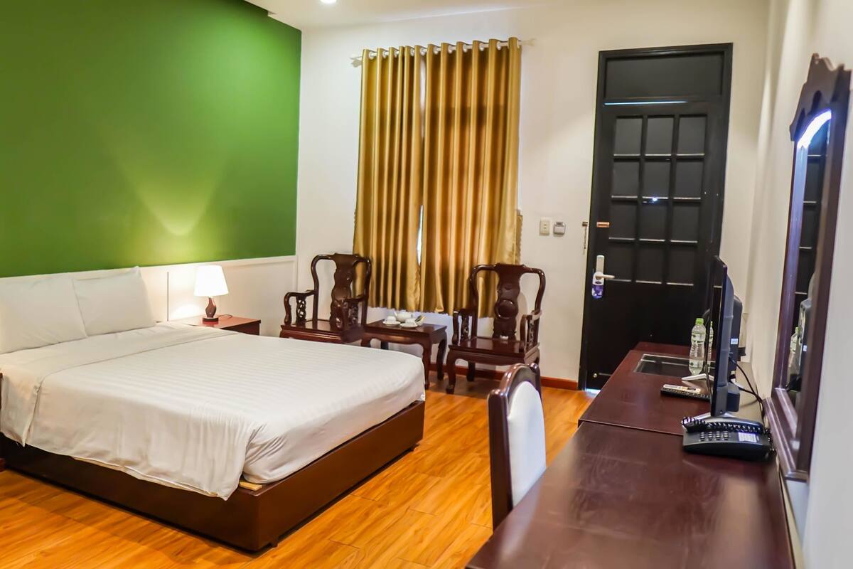 A hotel room with a large bed, two bedside tables with lamps, a wooden desk with a computer and a telephone, a chair, and a door with a window and curtains.