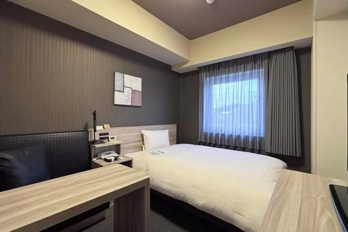 Ein Hotelzimmer mit Bett, Schreibtisch, Nachttisch, Lampe und einem Fenster mit Vorhängen.