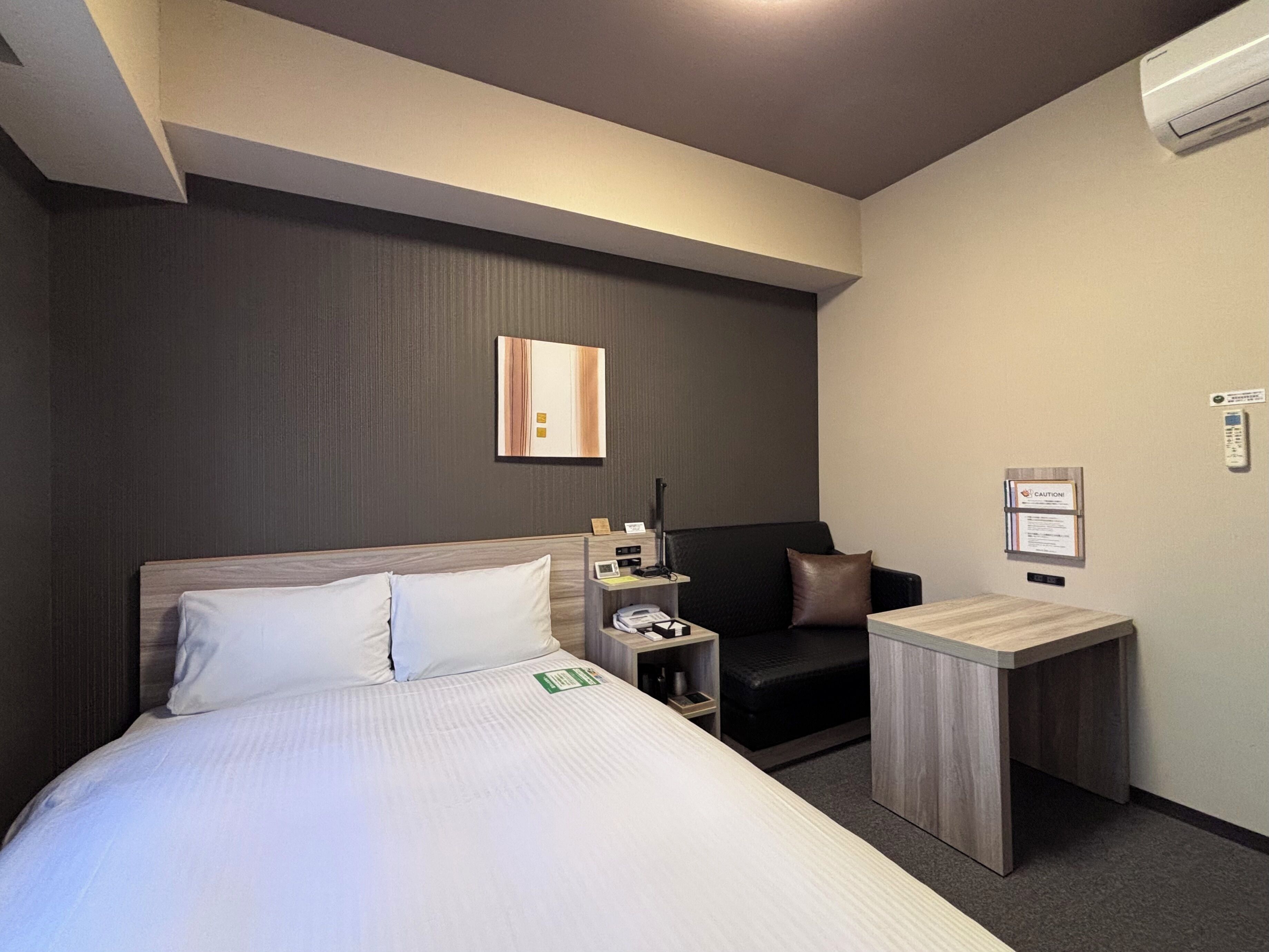 A hotel room with a bed, bedside table, chair, desk, and a wall-mounted air conditioner.