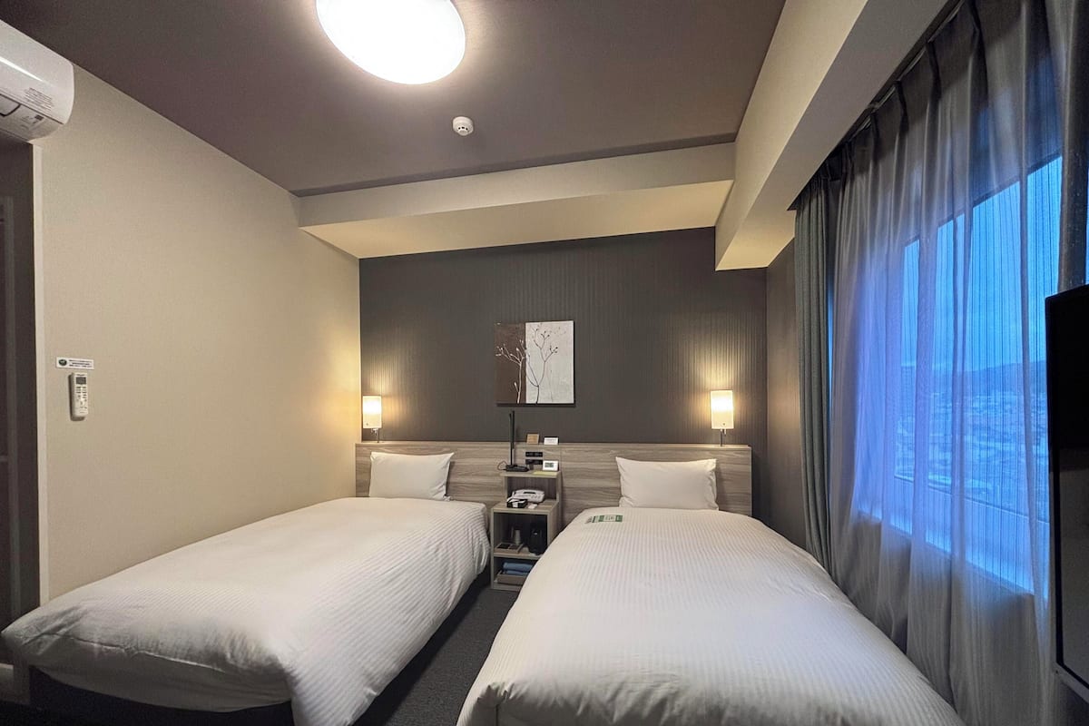 Ein Hotelzimmer mit zwei Einzelbetten, einer Deckenleuchte, einem an der Wand montierten Fernseher und einem Fenster mit Vorhängen.