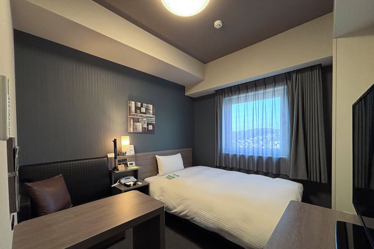 Ein Hotelzimmer mit Bett, Schreibtisch, Nachttisch, Lampe und Blick auf die Stadt durch das Fenster.