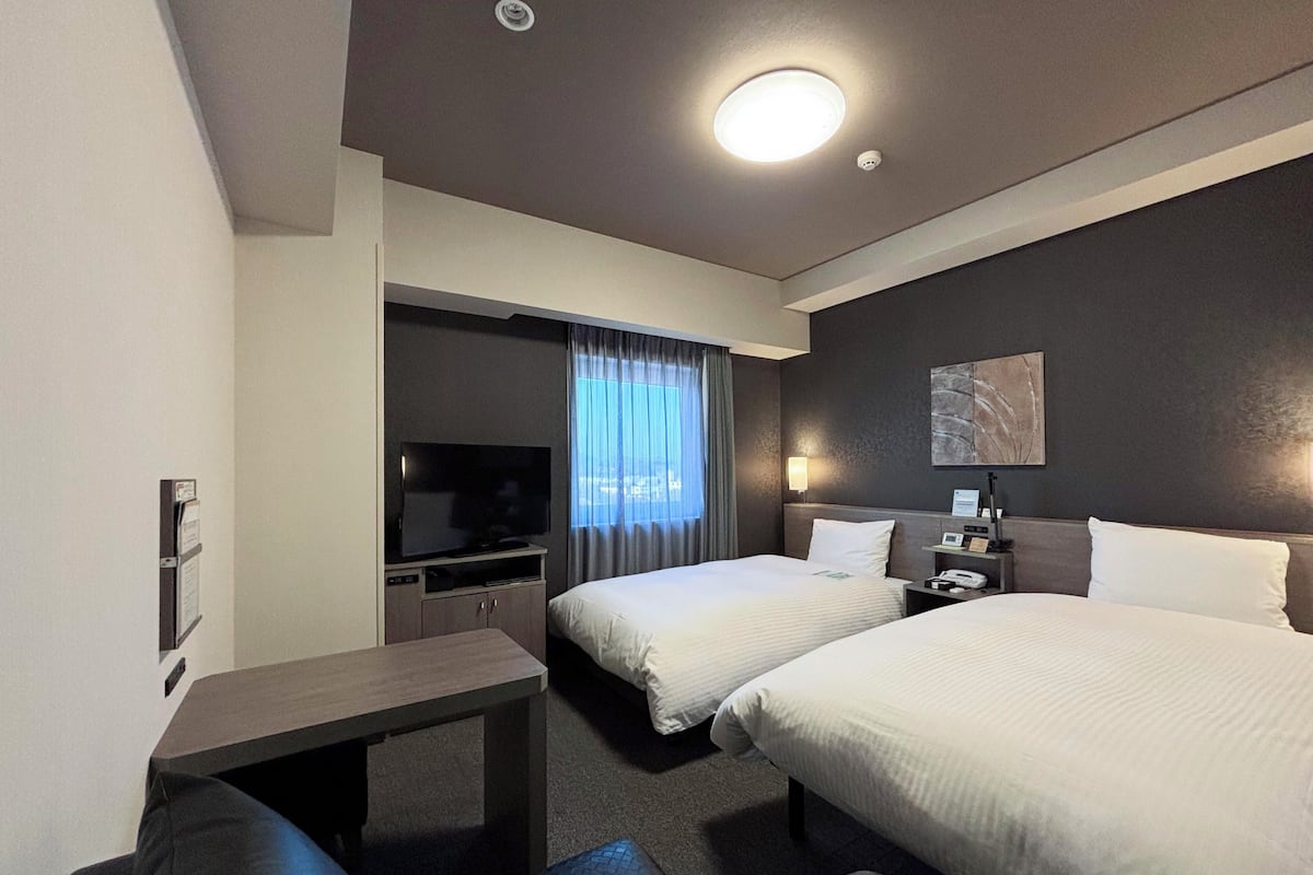 Ein Hotelzimmer mit zwei Betten, einem Schreibtisch, einem Fernseher, einem Nachttisch und einem Fenster mit Vorhängen.