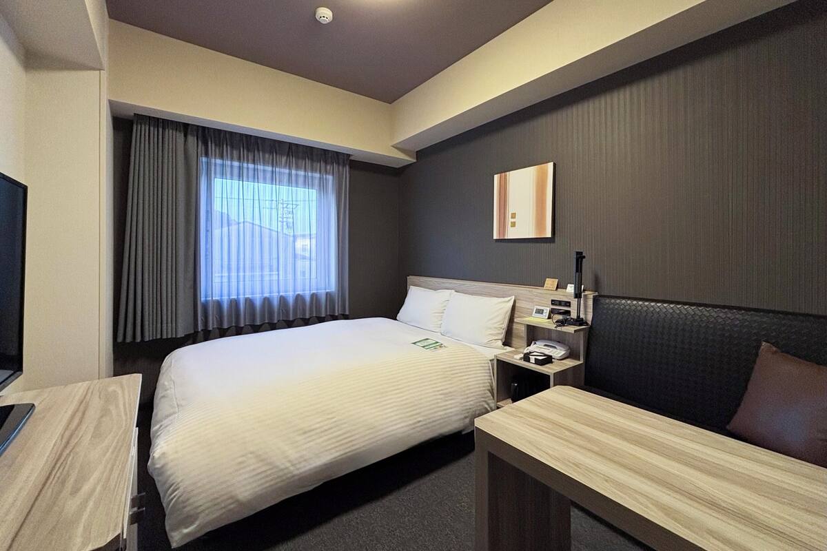 Ein Hotelzimmer mit Bett, Nachttisch, Lampe und Blick ins Freie durch ein Fenster.