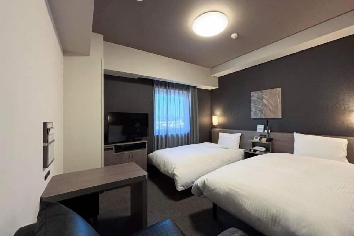 Ein Hotelzimmer mit zwei Betten, einem Schreibtisch, einem Fernseher und einem fensterbepflanzten Fenster.