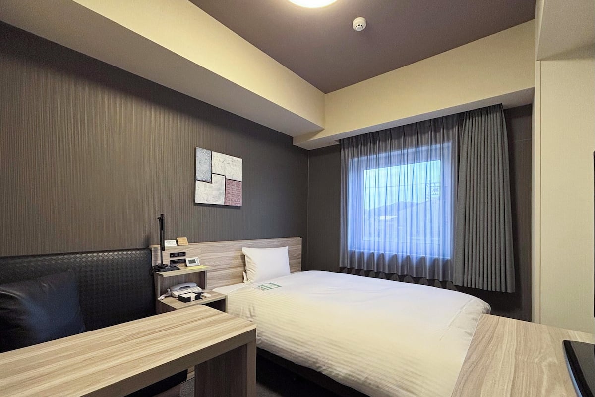 Ein Hotelzimmer mit Bett, Schreibtisch, Stuhl, Telefon und einem Fenster mit Vorhängen.