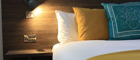 Una cama bien tendida con ropa de cama blanca y amarilla, un cojín estampado en color aguamarina y una lámpara de noche sobre una mesita de madera.