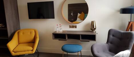 A hotel room with a bed, a yellow armchair, a grey armchair, a blue stool, a wooden desk with a mirror, a television, and a lamp.