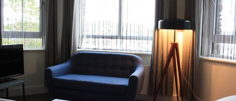 A hotel room with a blue sofa, a bedside table with a lamp, and a TV.