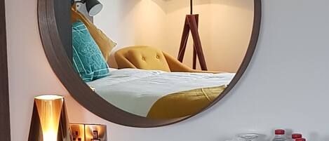 A round mirror with a wooden frame, a bedside lamp, a bed with yellow bedding, a floor lamp, and a minibar with bottled water and glasses.