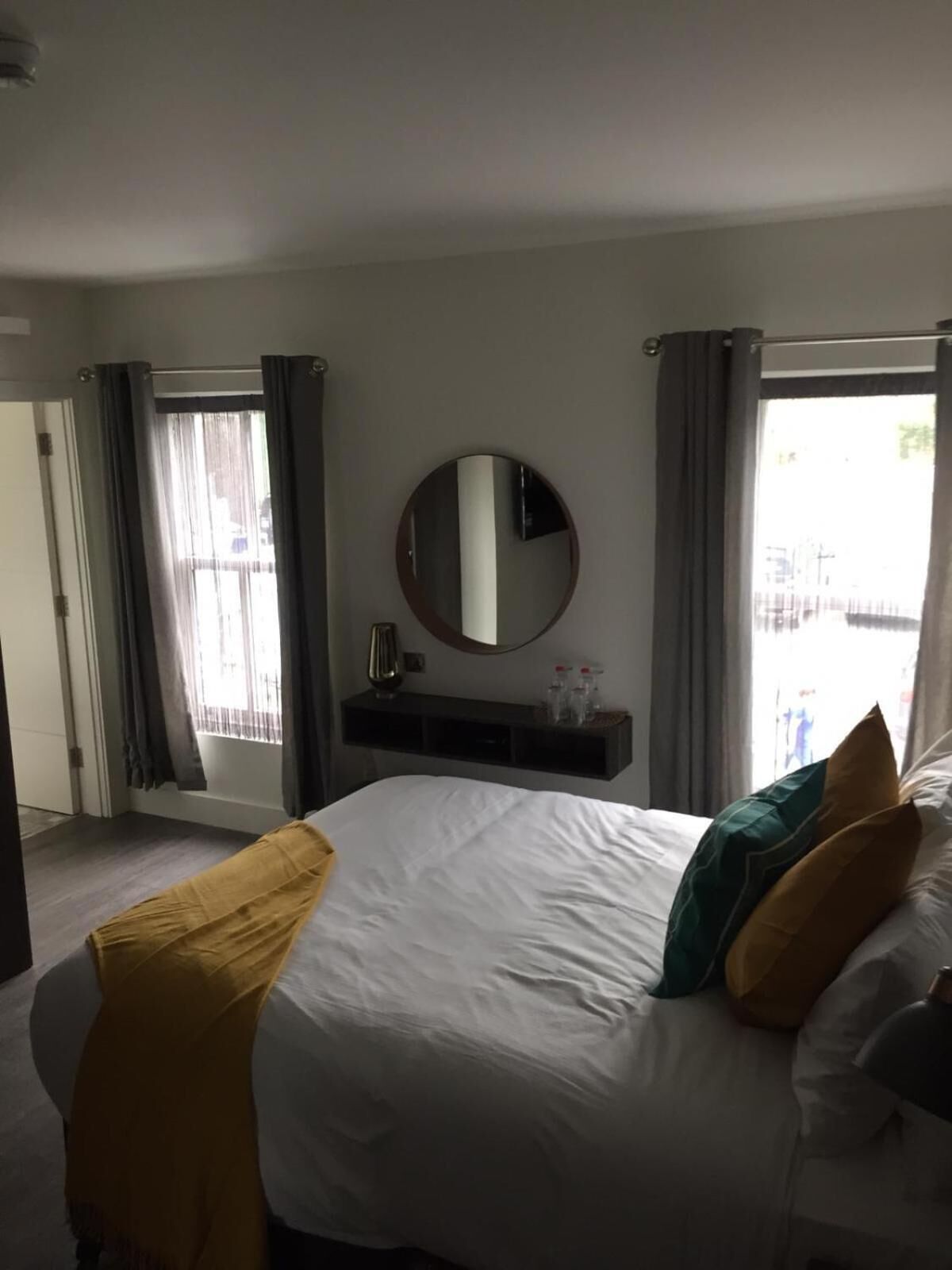 Una habitación de hotel con una cama grande, un espejo redondo, un estante pequeño con artículos de aseo y vistas por la ventana.