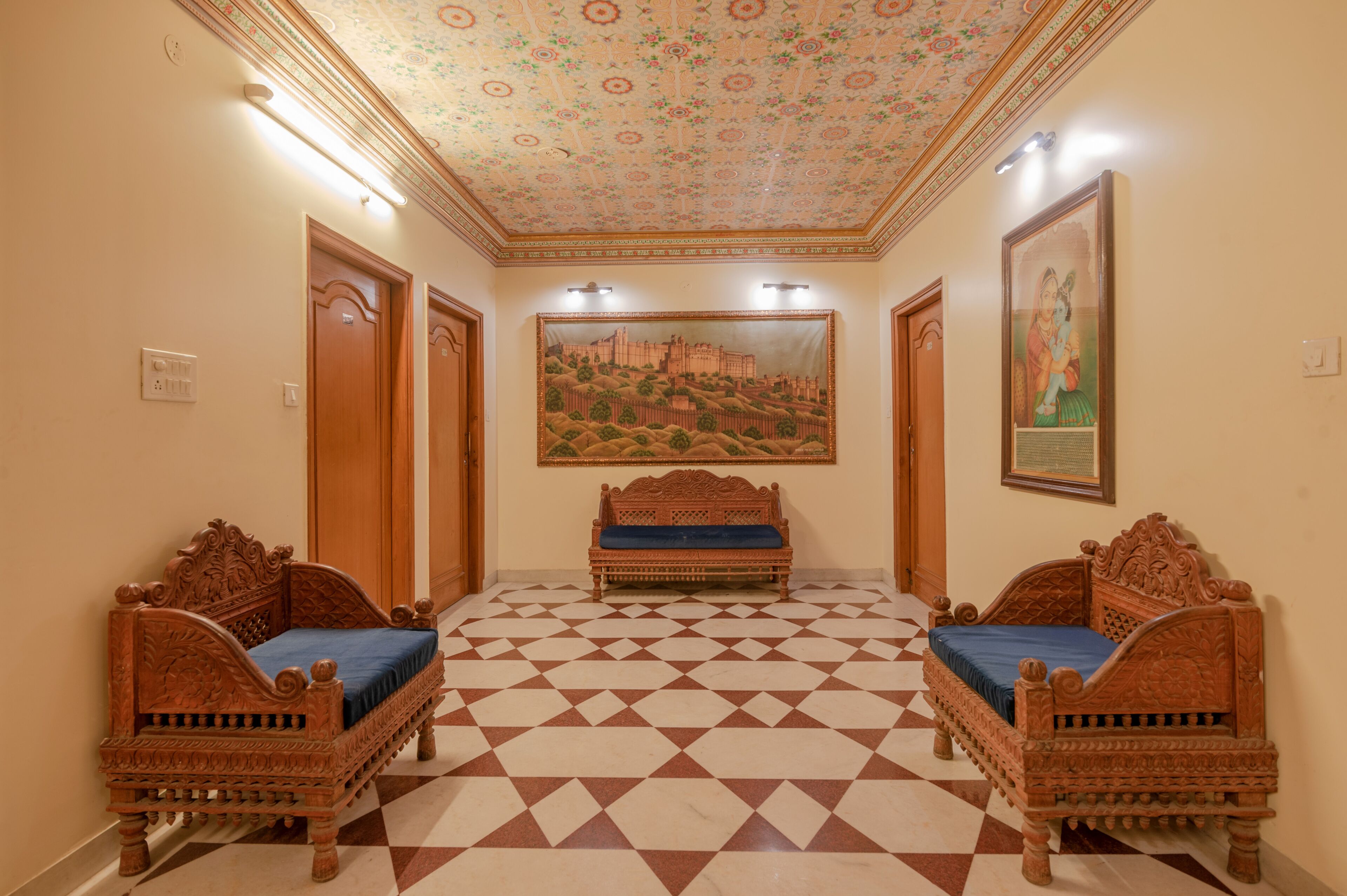 A hallway with a checkered floor, two ornate wooden beds with blue cushions, a large tapestry on the wall, and a ceiling with a floral pattern.