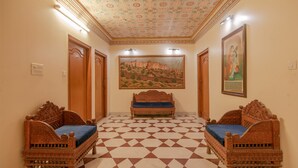 Un couloir doté d’un sol à carreaux, deux lits en bois richement décorés avec des coussins bleus, une grande tapisserie au mur et un plafond orné de motifs floraux.