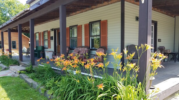 A covered porch with wooden columns, a seating area with chairs, and a garden with flowers. - RIVERVIEW ADIRONDACK RUSTIC APARTMENT (Warrensburg)