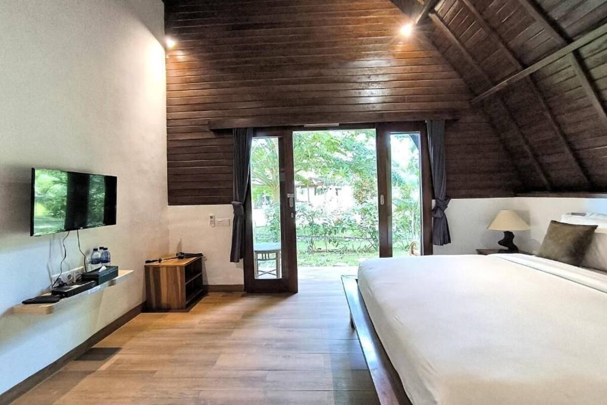 Ein Schlafzimmer mit einem großen Bett, einem an der Wand montierten Fernseher, einer Holzzieldecke und Blick ins Grüne.